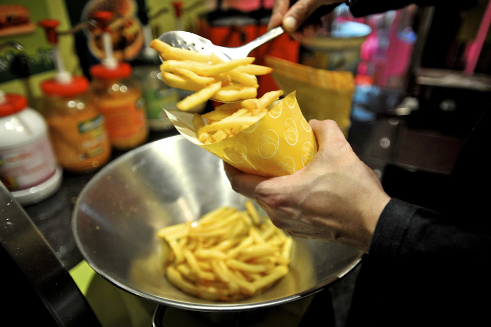 A person using a slotted spoon to fill a yellow cone with French fries from a metal bowl. Various condiments are in the background.