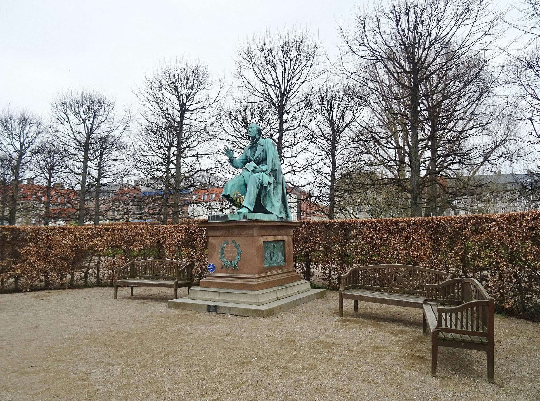 정원을 배경으로 나무와 벤치로 둘러싸인 받침대에 앉은 남자의 청동 동상.