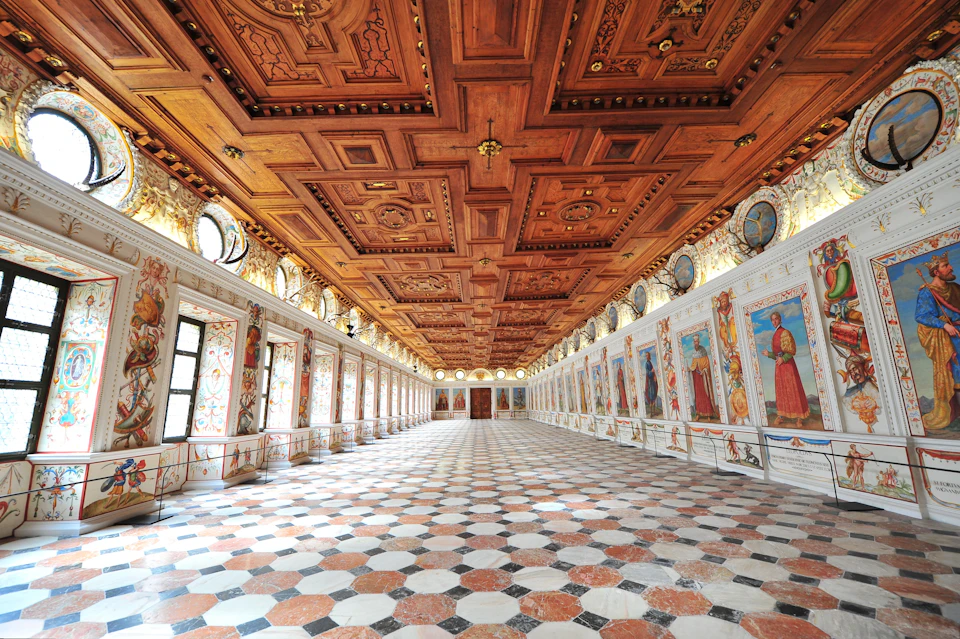 A grand hall with ornate painted walls, intricate wooden ceiling, and a checkered floor, lined with tall windows.