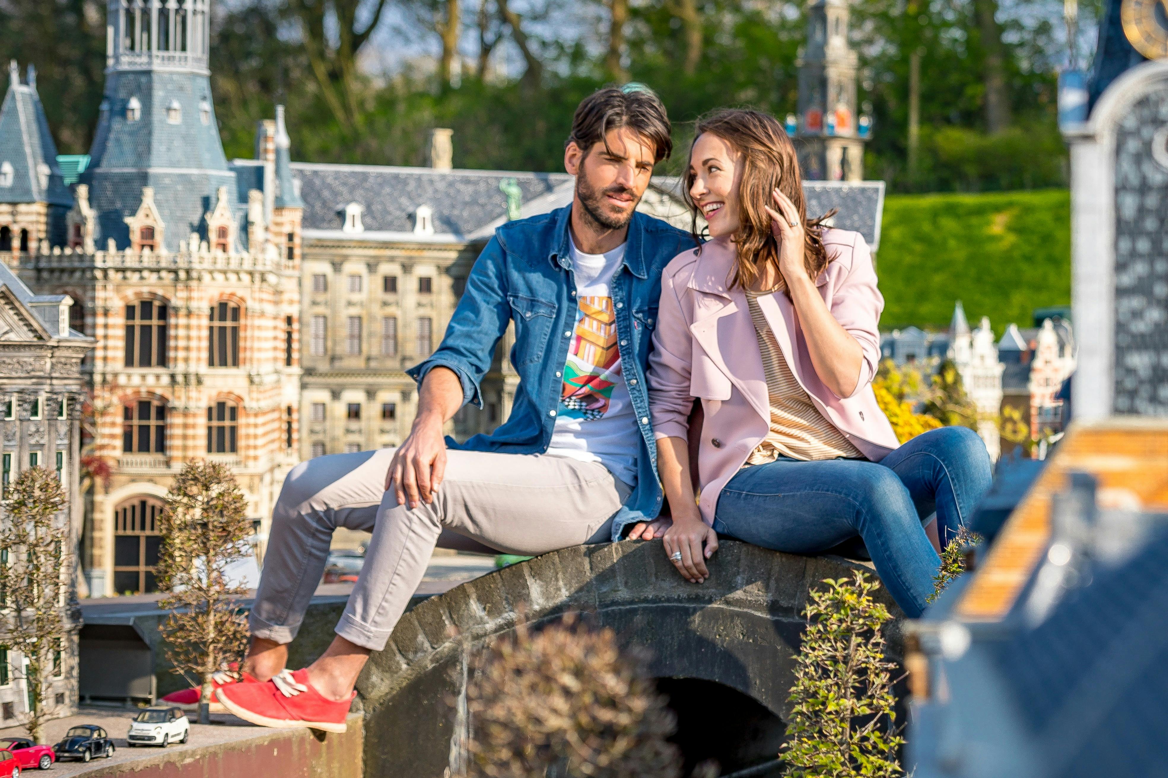 공원의 미니어처 건물과 녹지를 배경으로 작은 다리에 앉아 있는 남성과 여성.