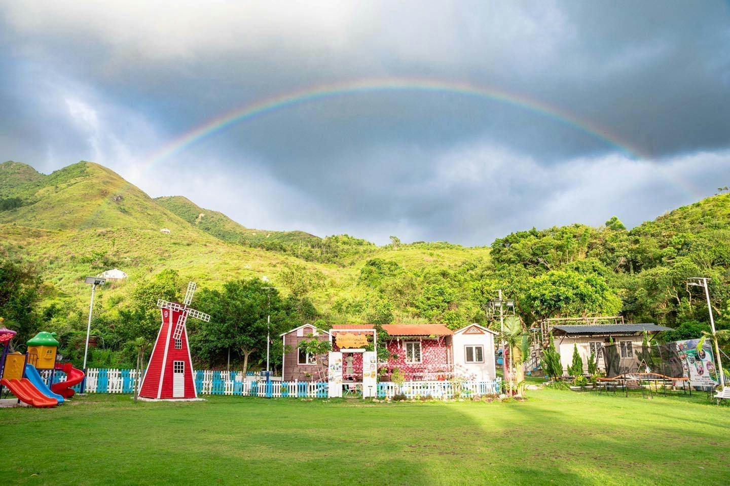 A colorful scene featuring a small red windmill and quaint houses with a rainbow arching over lush green hills.
