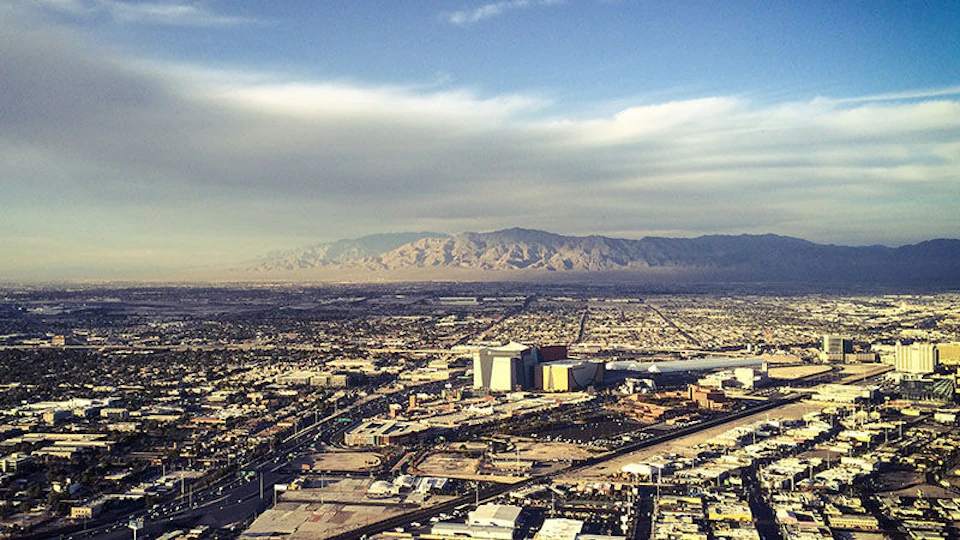 Aerial view of a sprawling cityscape with modern buildings, roads, and vast mountains under a partly cloudy sky.