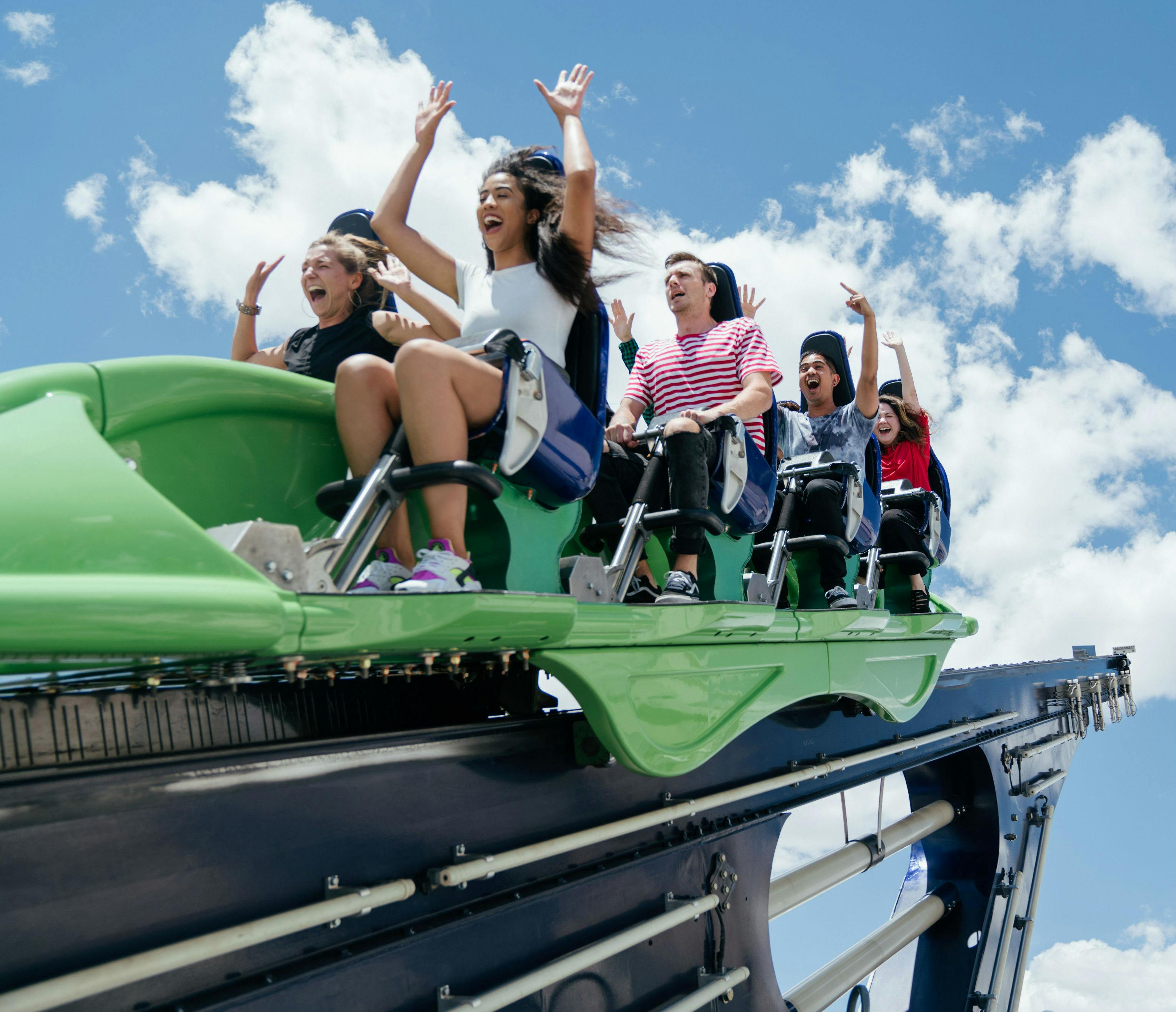 Ludzie jeżdżący zielono-czarną kolejką górską, podnosząc ręce i krzycząc z radości na błękitnym niebie z chmurami.