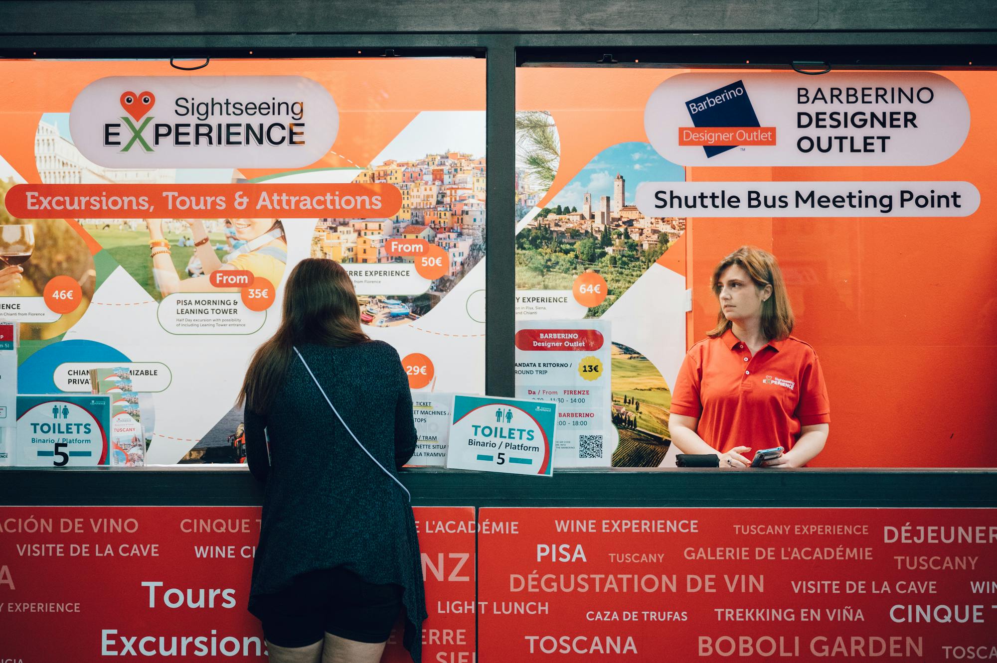 一位身穿红色Polo衫的女士站在Barberino Designer Outlet展台前的办公桌前，这是一个繁忙的穿梭巴士集合点。