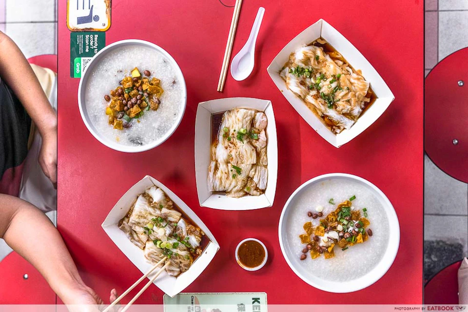 Top-down view of a red table with bowls of congee, plates of rice rolls, a pair of chopsticks, a spoon, and a small sauce dish.