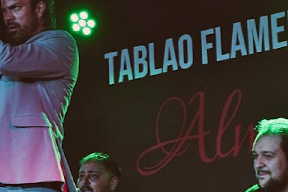 A flamenco dancer performs on stage, accompanied by a guitarist and a singer, with a backdrop reading "Tablao Flamenco."