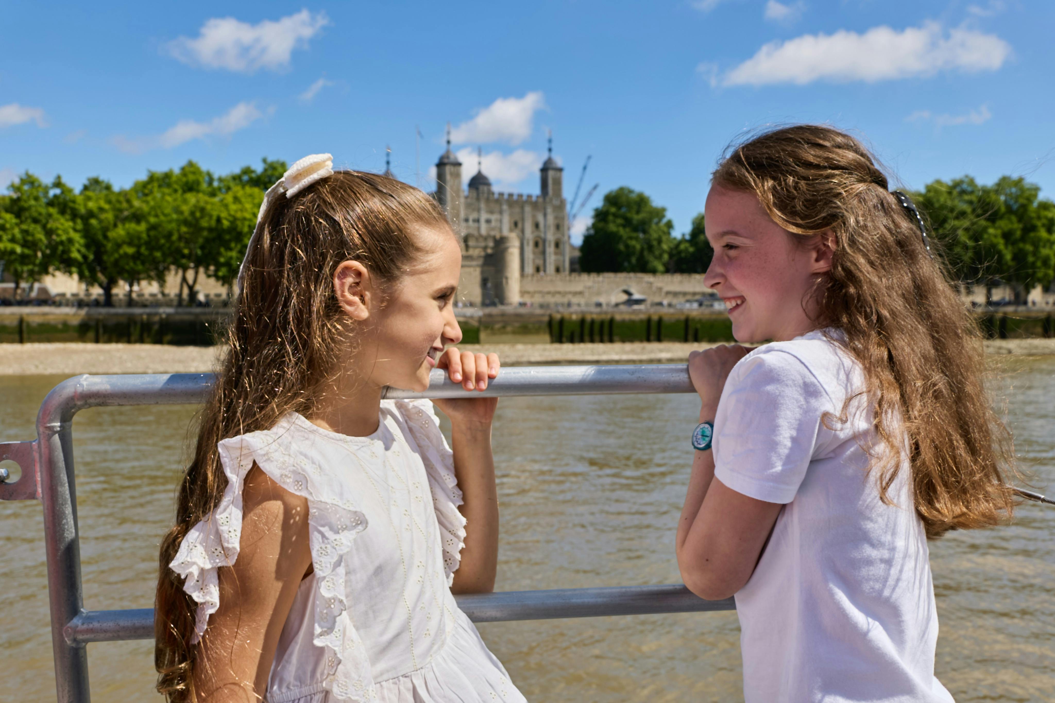 Deux jeunes filles vêtues de blanc qui se sourient sur un bateau, avec un château historique et une rivière en arrière-plan.