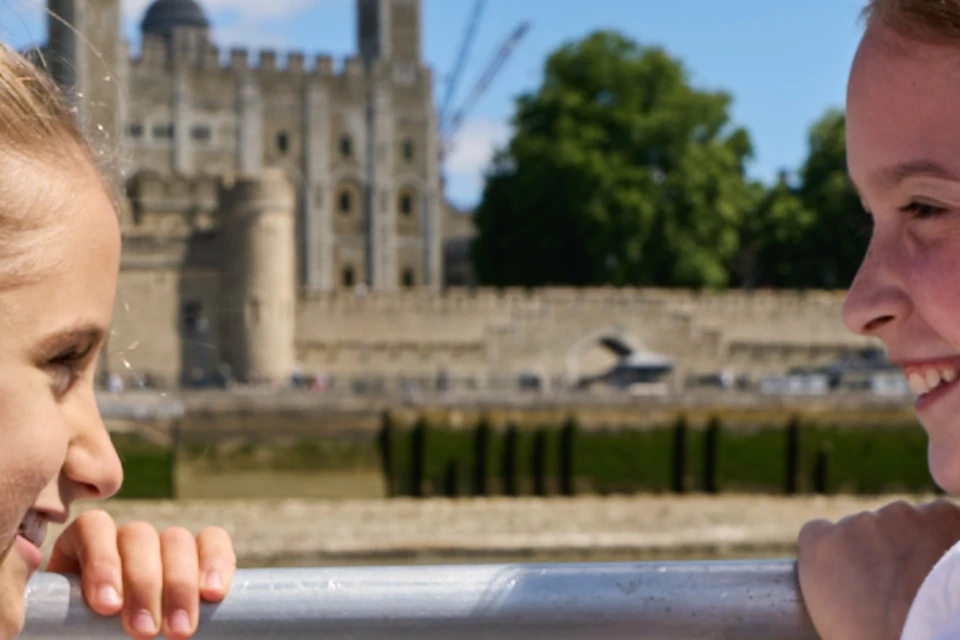 Two girls in white outfits smiling at each other on a boat, with a historic castle and river in the background.
