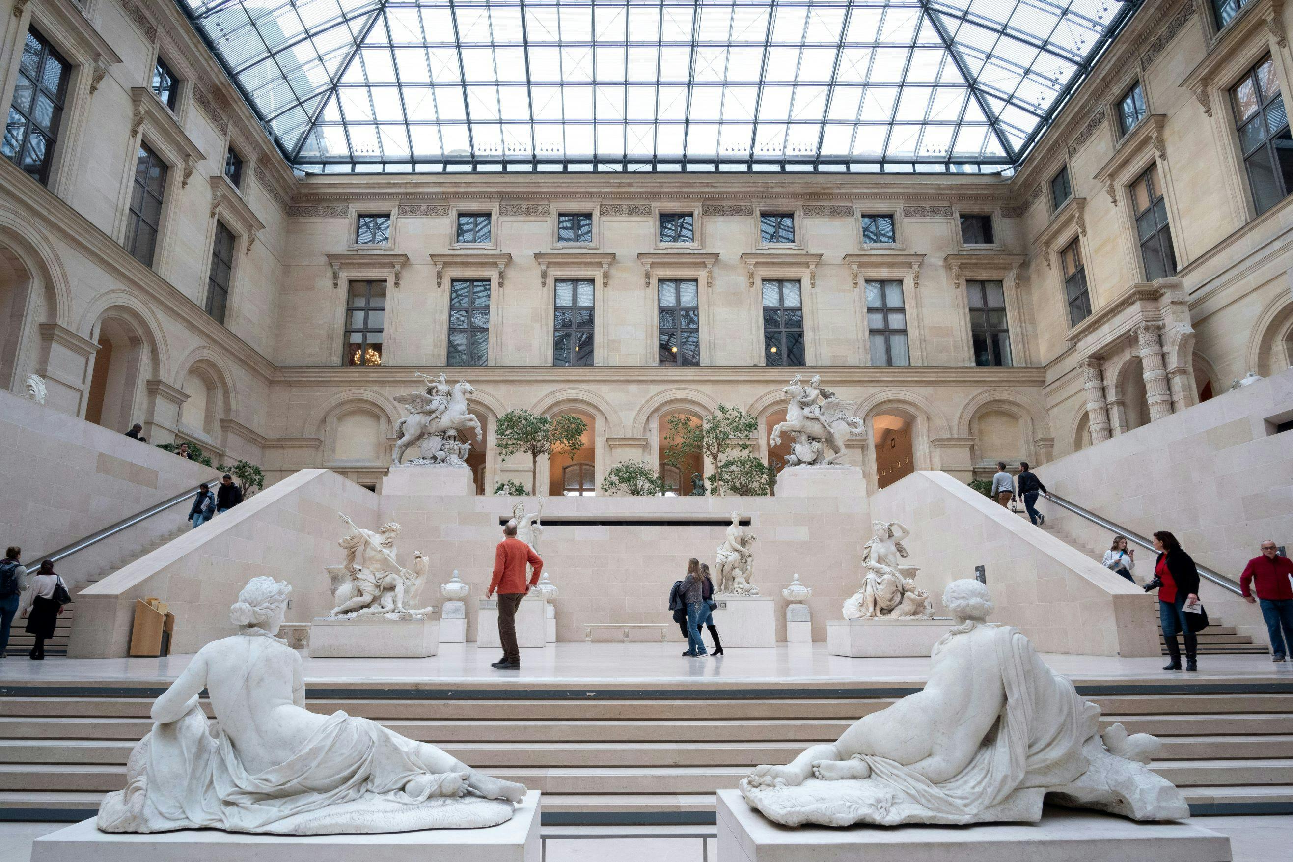 Une salle de musée avec des sculptures, des escaliers et un plafond de verre, où les visiteurs se promènent et admirent les objets exposés.