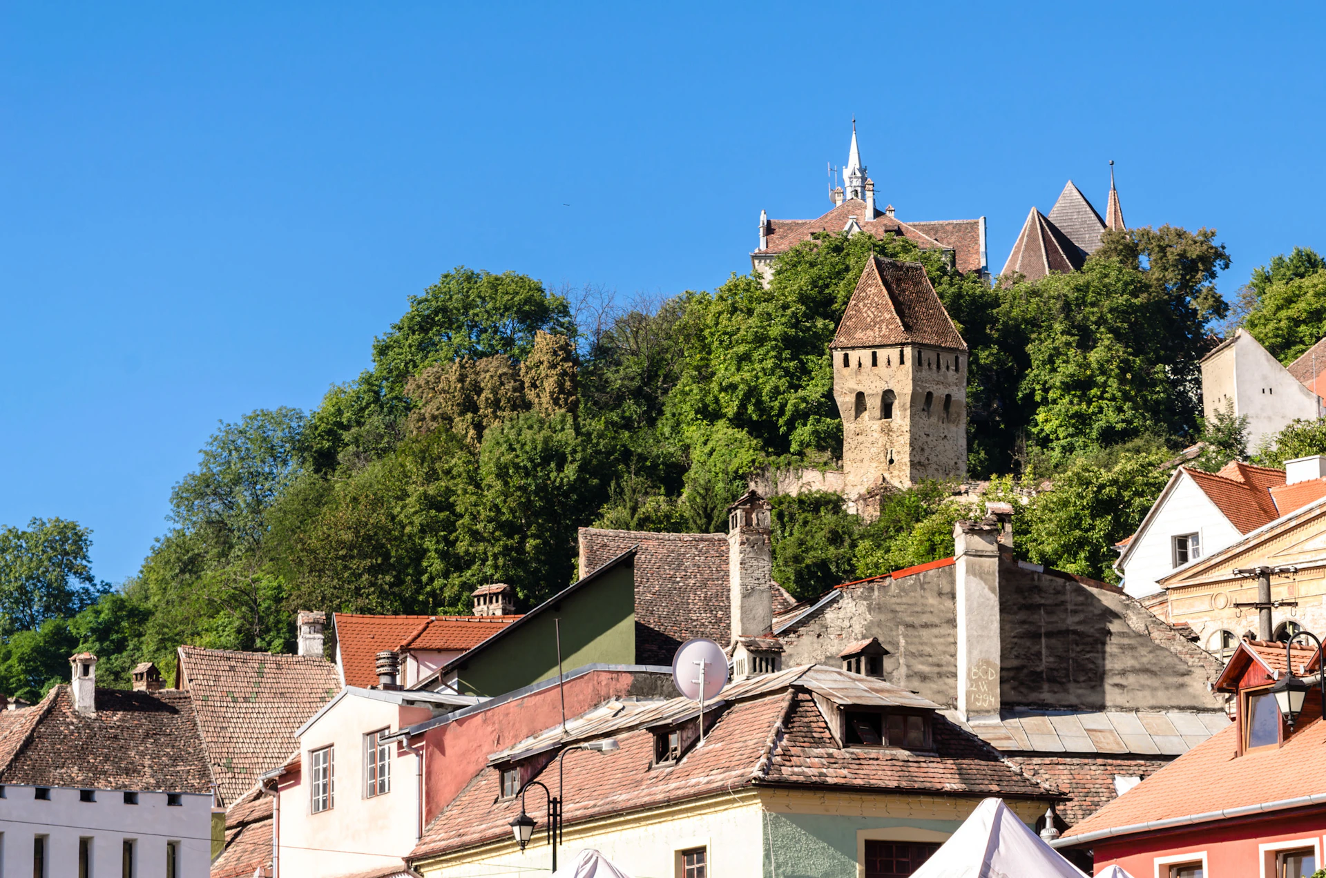 Colorful houses with tiled roofs at the base of a hill, topped with a tower and an old church, surrounded by dense greenery and a clear blue sky.