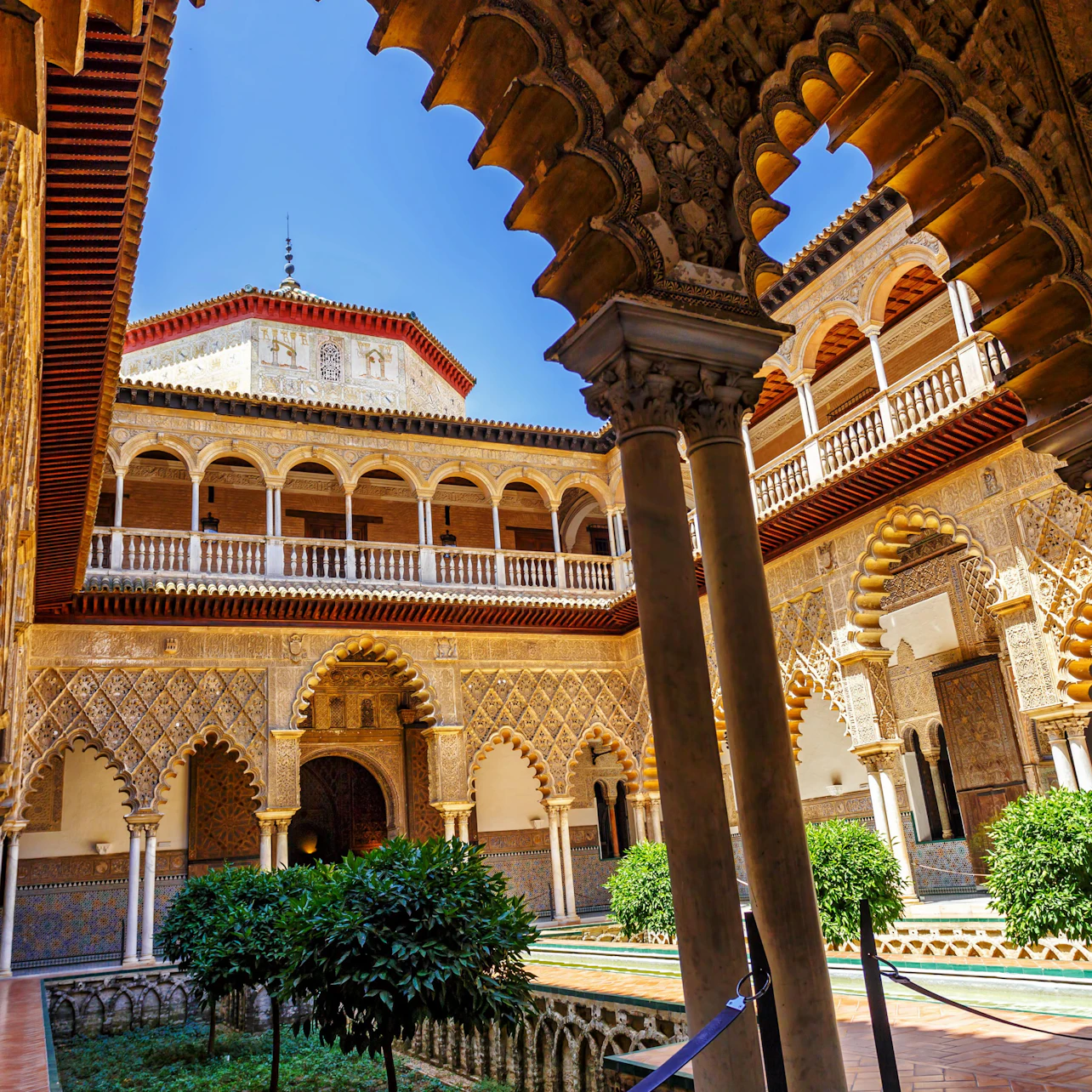 A historic courtyard with intricate arches, columns, a central pool, and lush greenery under a clear blue sky.