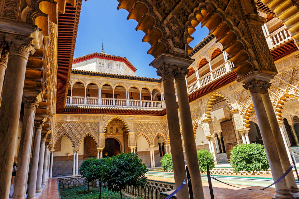 A historic courtyard with intricate arches, columns, a central pool, and lush greenery under a clear blue sky.