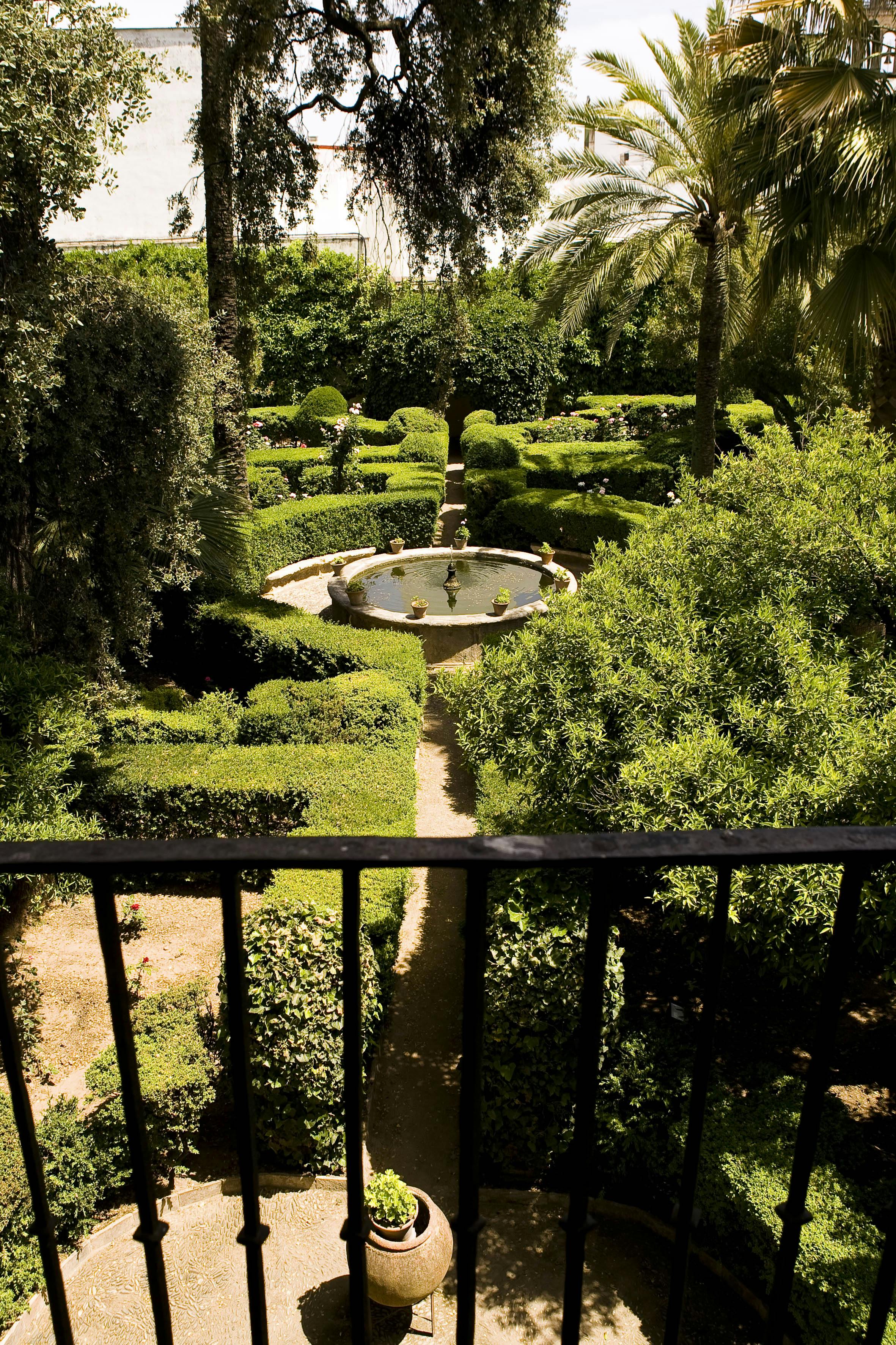 A formal garden with manicured hedges, a fountain at the center, and a pathway visible from a wrought iron balcony.