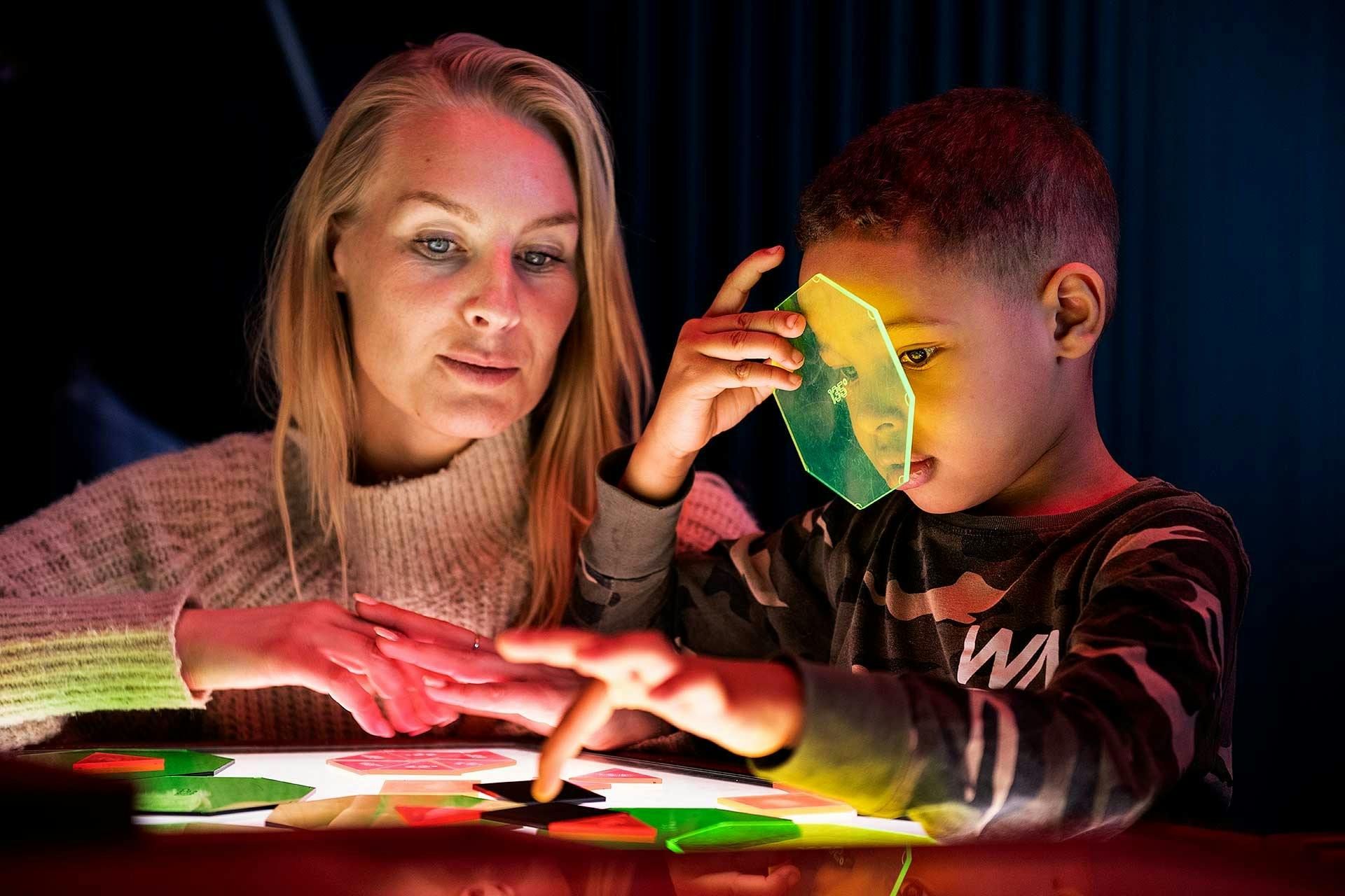 Žena a dítě interagují s barevnými průsvitnými tvary na světelném stole, přičemž dítě drží u oka zelený tvar.