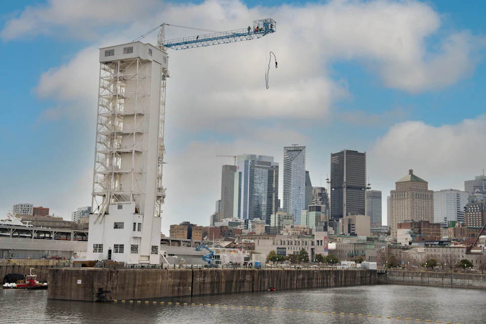A crane and a tall white structure by a waterfront, with a city skyline in the background under a partly cloudy sky.