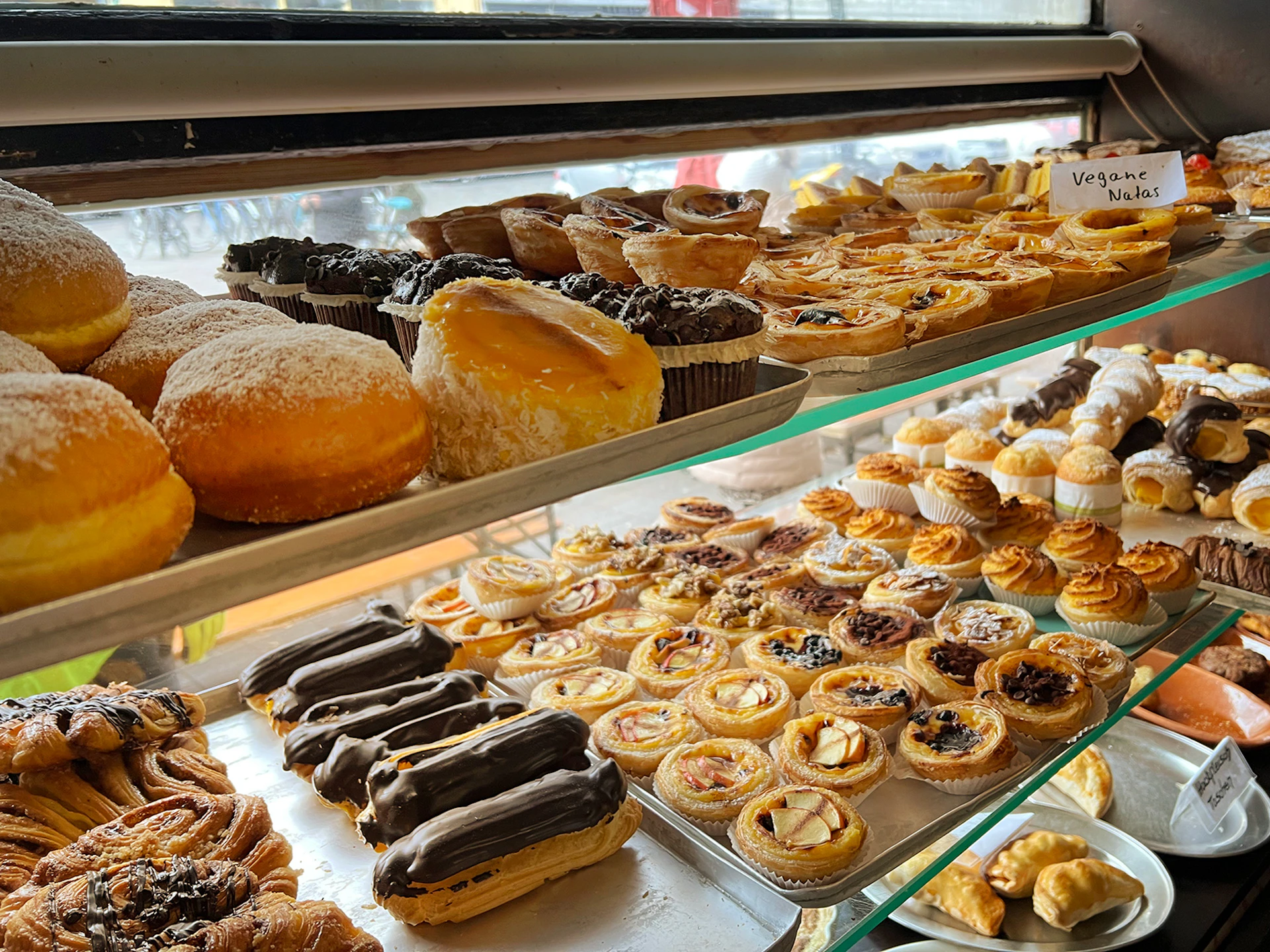 A variety of pastries including eclairs, tarts, and donuts displayed on metal and glass shelves in a bakery.