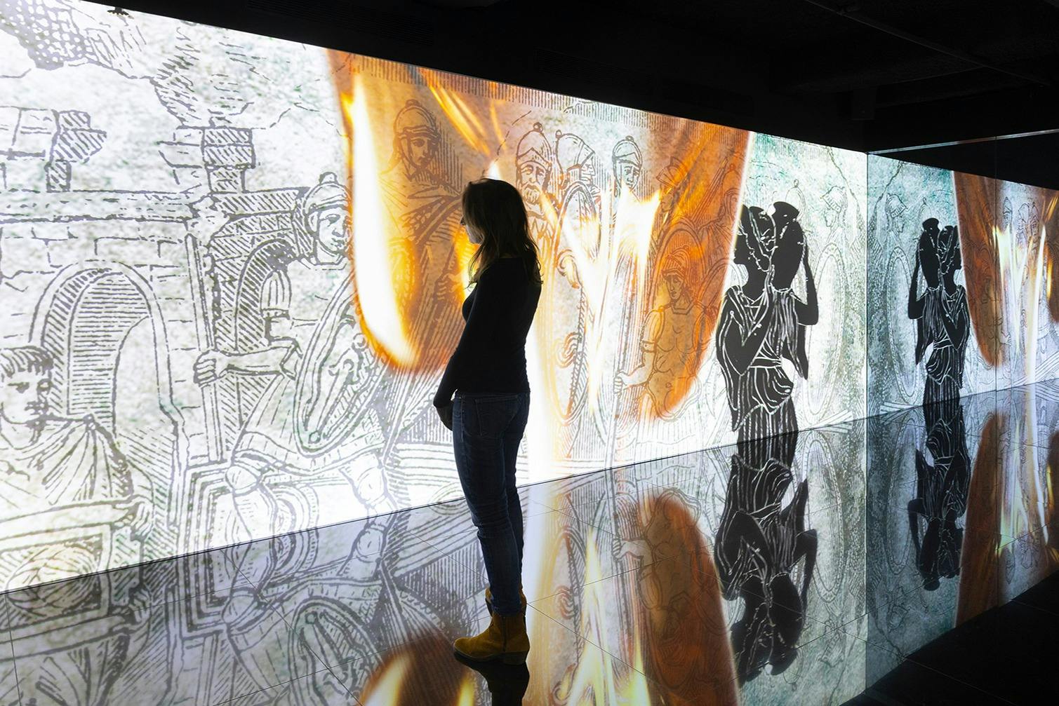 Osoba stojí před osvětlenou zdí s rytinami starověkých válečníků a reflexní podlahou.