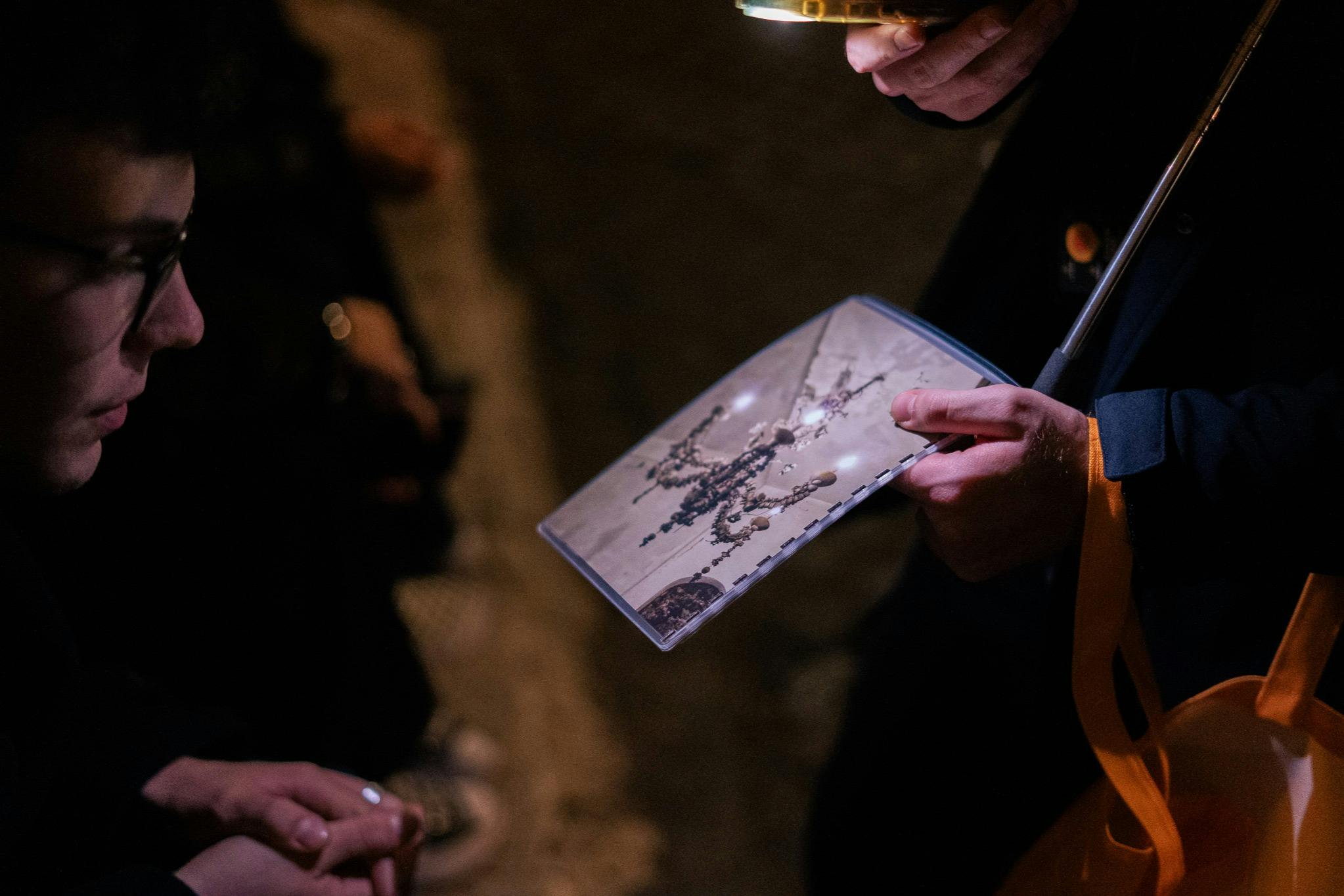 A person holding a booklet displaying jewelry designs under dim lighting, surrounded by blurred figures.