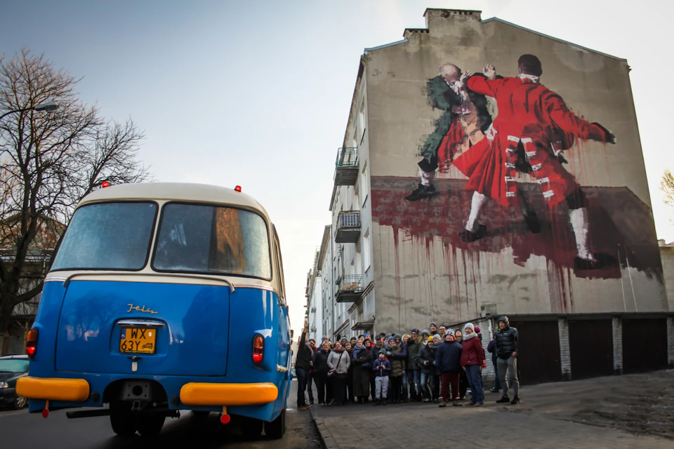 A crowd stands near a blue and white van, in front of a tall building featuring large, colorful mural art.