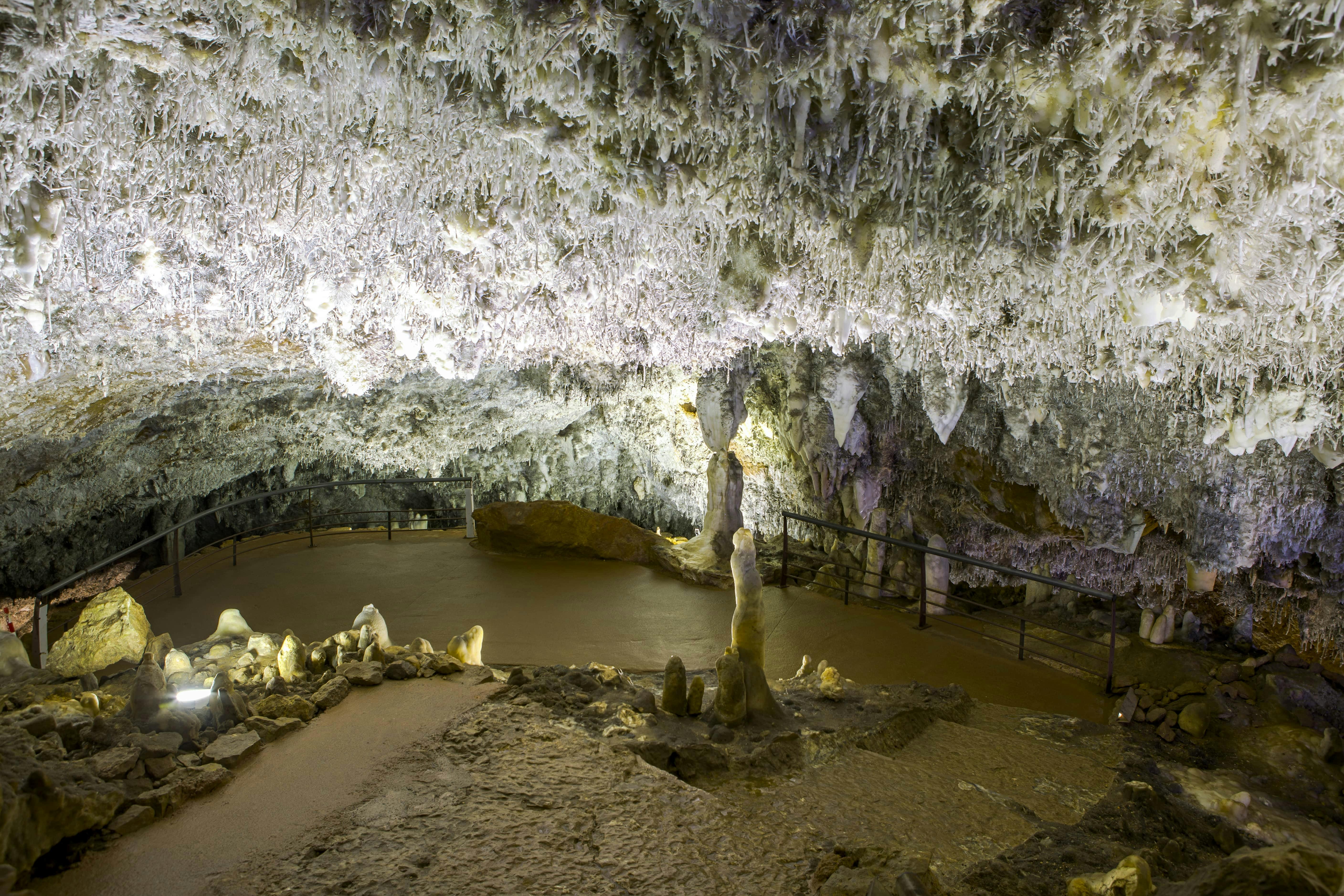 Huleinteriør med takkede stalaktitter i loftet, stalagmitter på gulvet og en metalgangbro med rækværk.