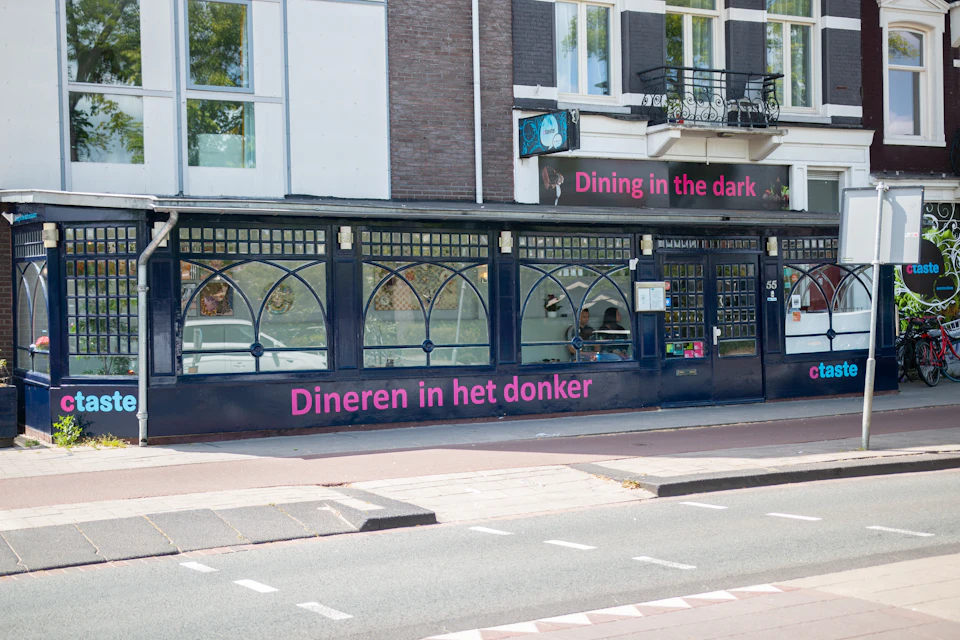Restaurant exterior with signs "Dining in the dark" and "Dineren in het donker" in pink lettering.