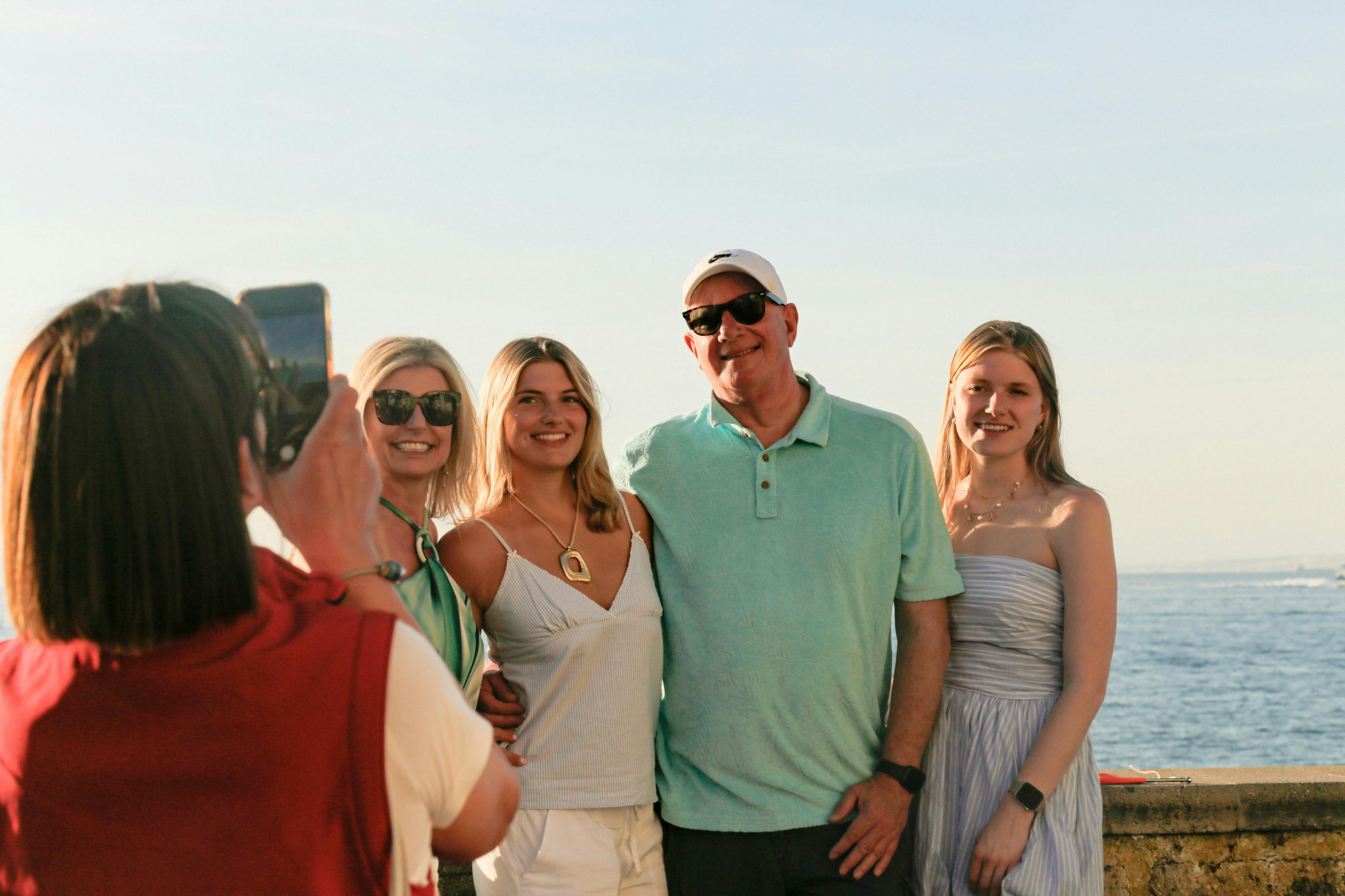 Drie mensen staan op een zonnige dag voor een watermassa; de man in het midden draagt een groen shirt en een witte pet.