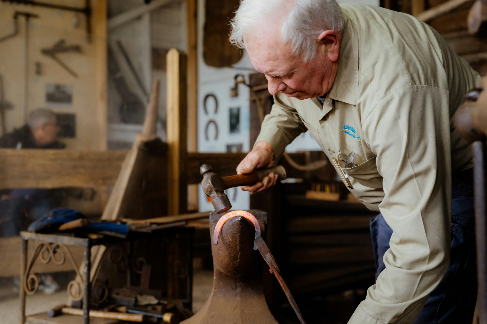 An older man wearing a beige shirt uses a hammer on a horseshoe placed on an anvil in a workshop.