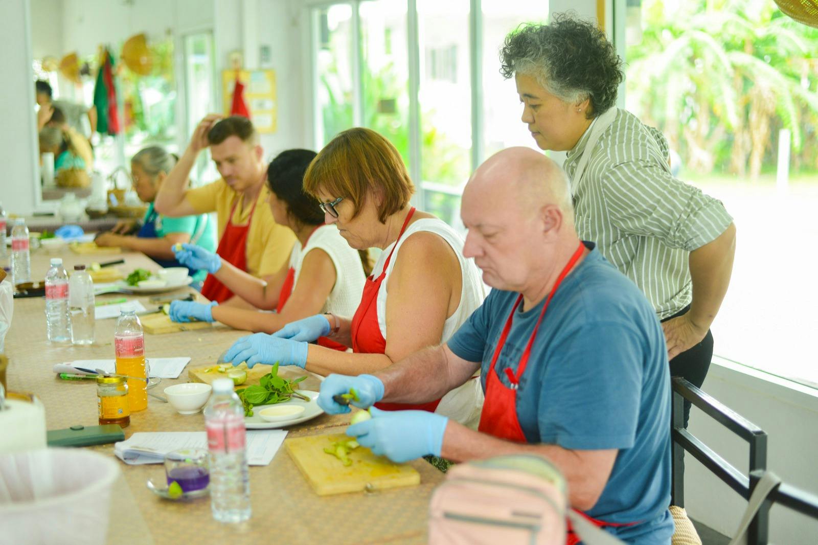 一群穿着红色围裙和蓝色手套的人正在餐桌上准备食物，由一名妇女监督。