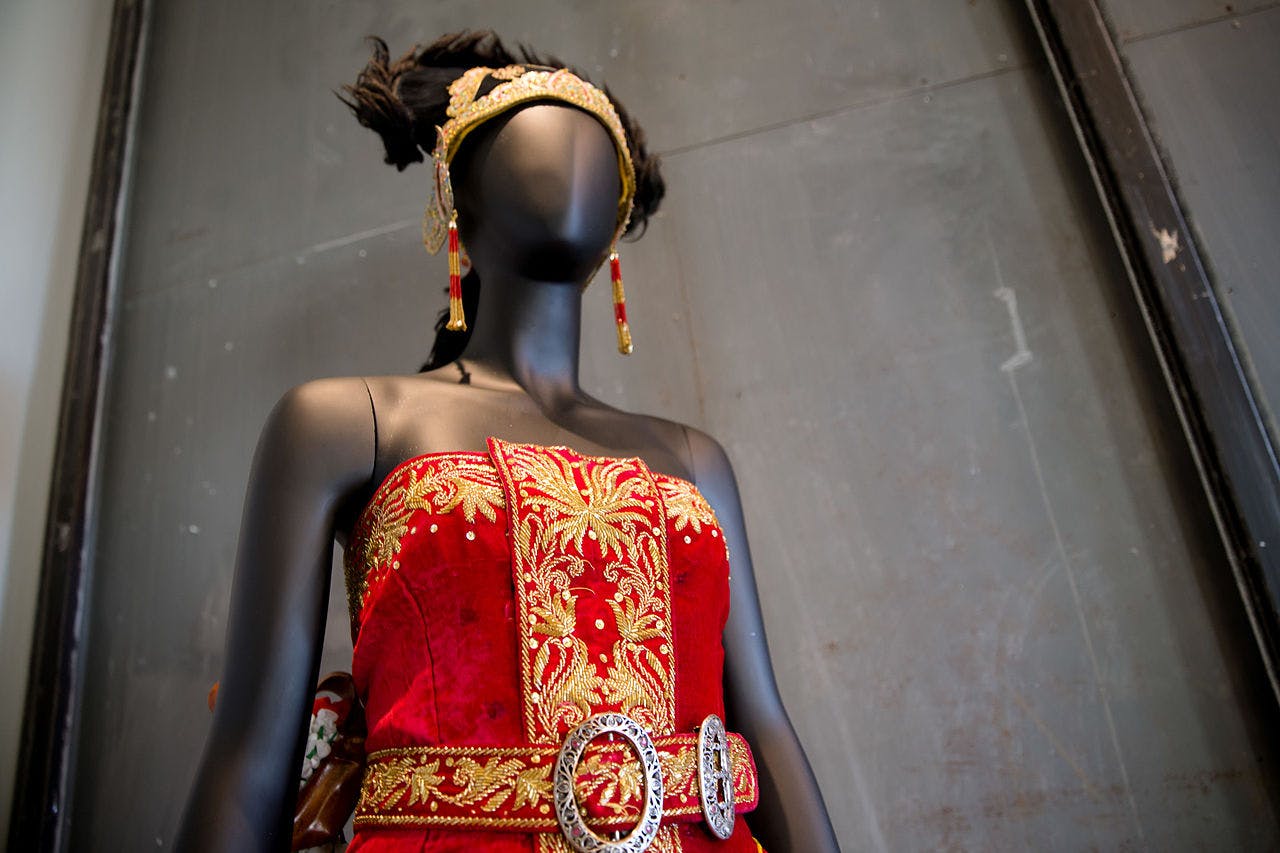 A mannequin wearing a red and gold ornate dress with matching headdress and earrings, displayed in front of a plain wall.