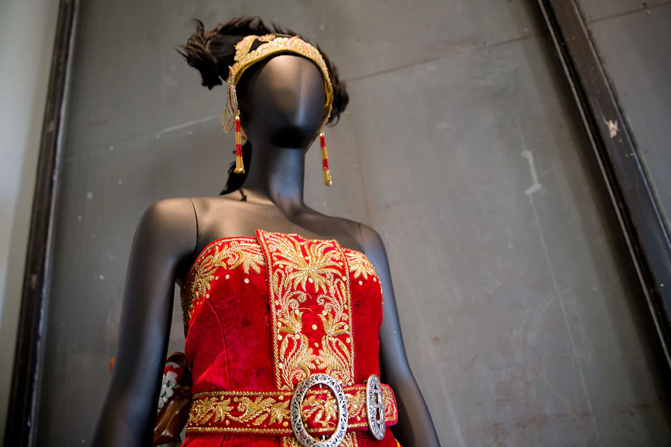 A mannequin wearing a red and gold ornate dress with matching headdress and earrings, displayed in front of a plain wall.