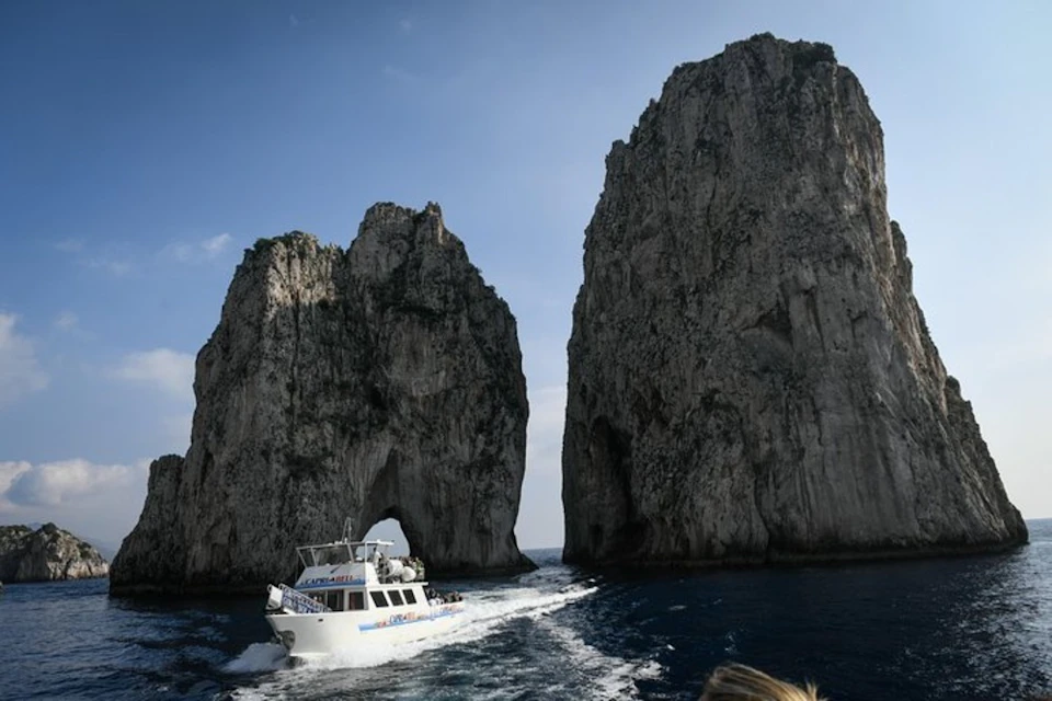 A small boat cruises between two large, rugged rock formations in the sea under a clear sky.