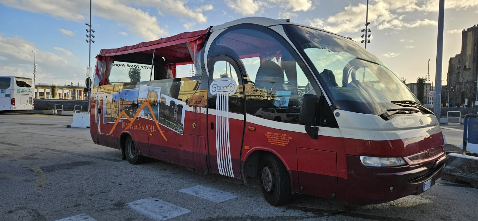 A red sightseeing bus with large side windows and promotional images of Naples landmarks parked in a lot under a cloudy sky.