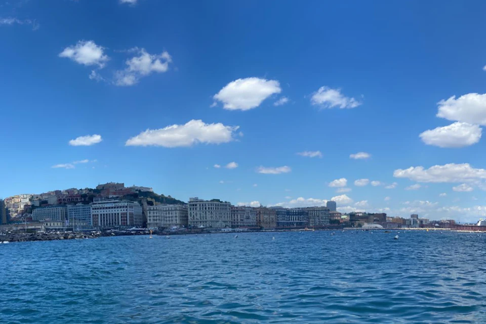 A waterfront cityscape under a clear blue sky with scattered clouds, featuring buildings along the shoreline and calm ocean waters.