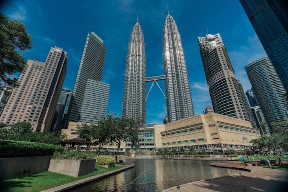 스카이브리지로 연결된 쌍둥이 빌딩을 포함한 높은 고층 건물이 있는 도시 풍경으로, 전면에 공원과 연못이 있습니다.