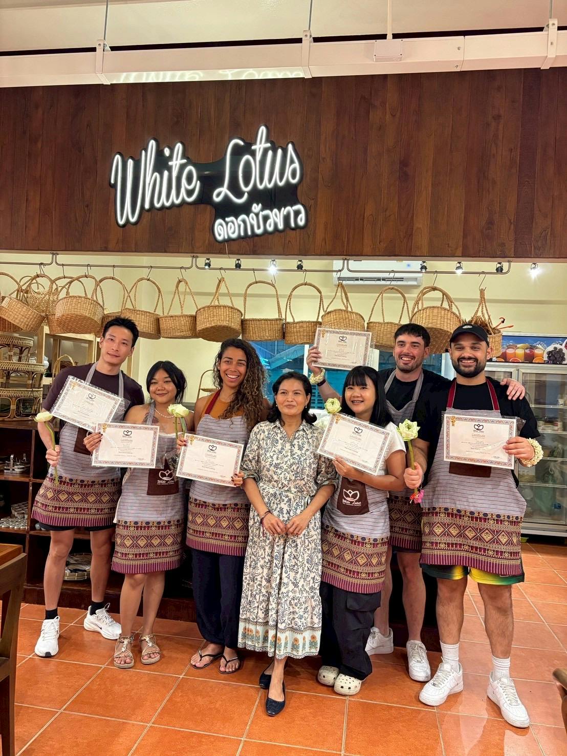 A group of people holding certificates and flowers, standing in front of a sign reading 'White Lotus,' with baskets displayed above.