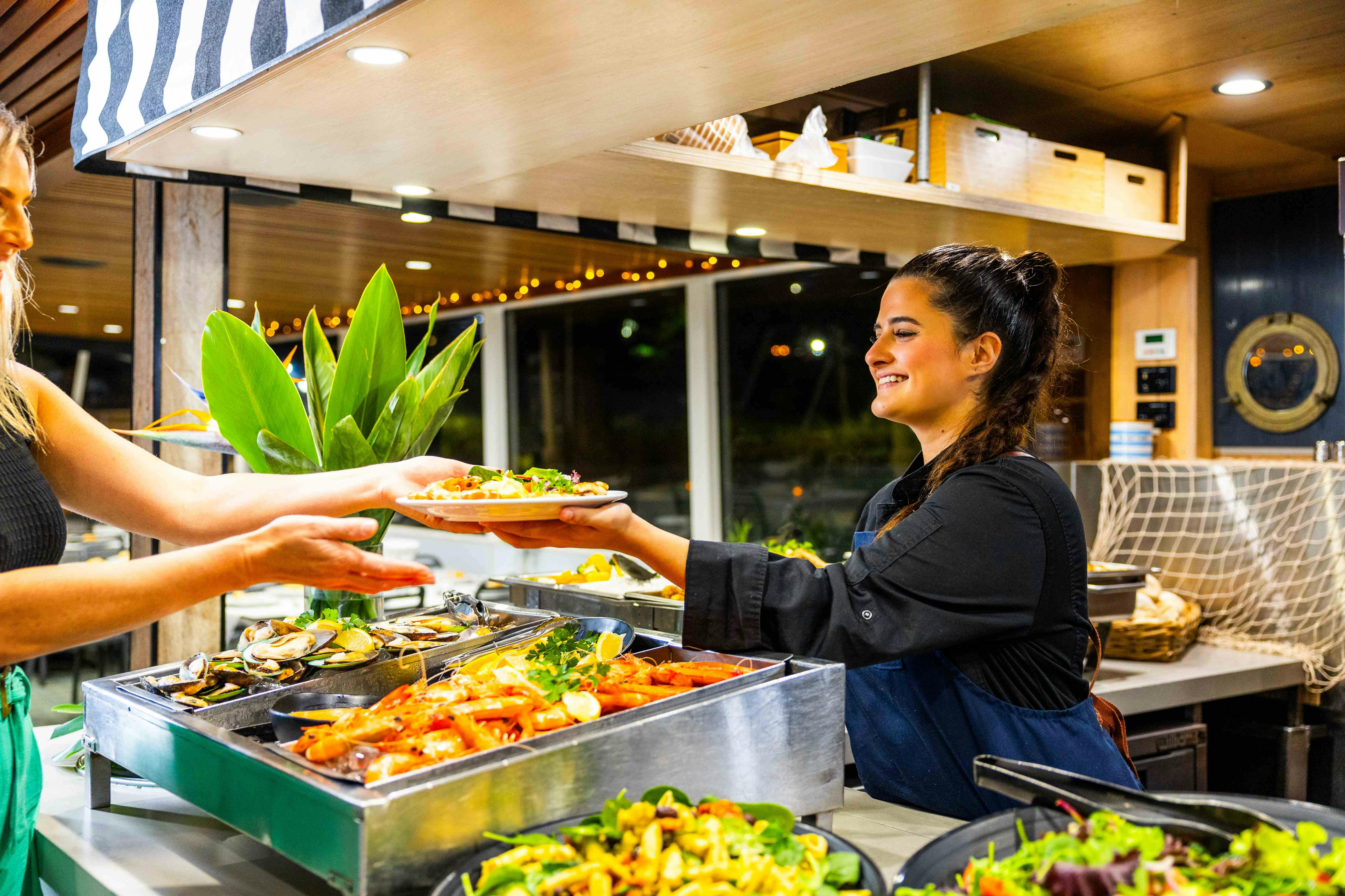 一位身穿黑色制服的微笑女士在餐厅的沙拉吧提供食物，餐厅装饰为木制，灯光明亮。
