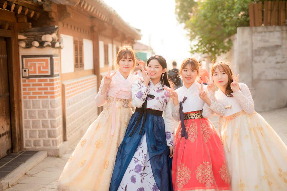 Four women wearing traditional Korean hanboks stand together, smiling and making peace signs in a sunlit outdoor setting.