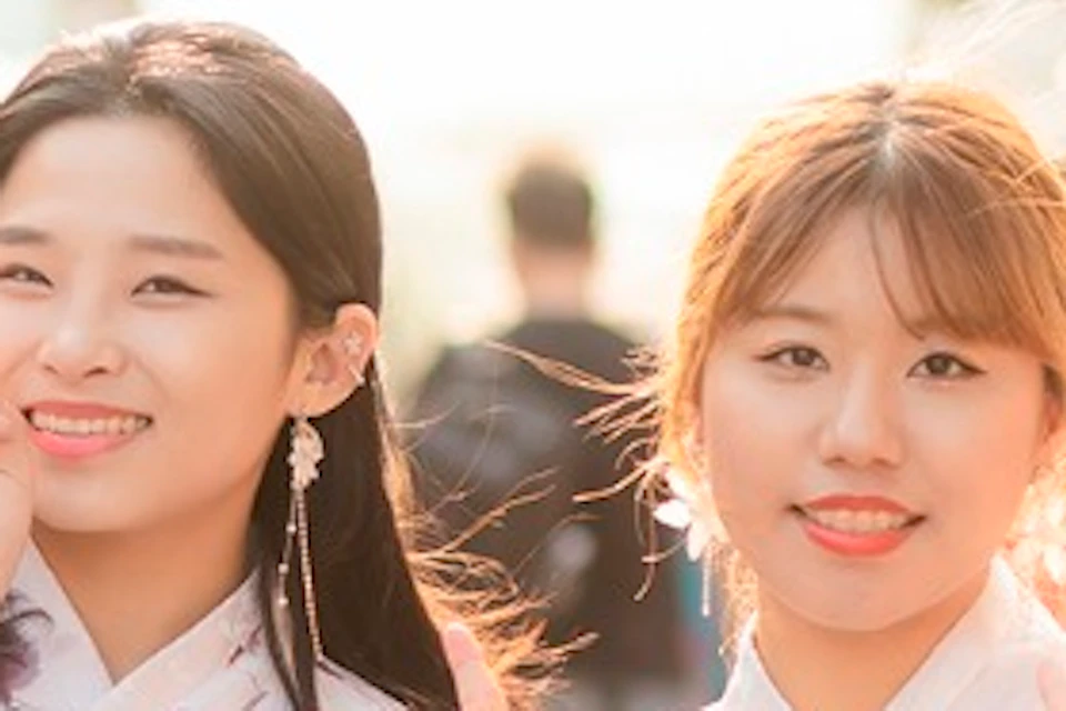 Four women wearing traditional Korean hanboks stand together, smiling and making peace signs in a sunlit outdoor setting.