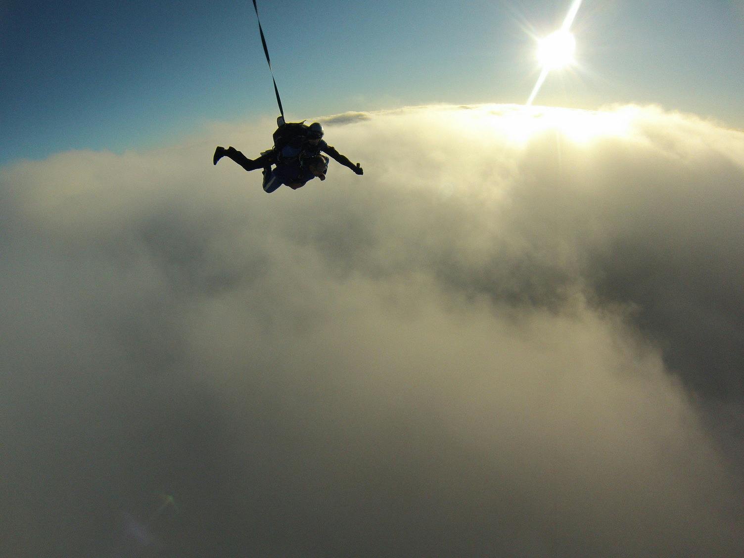 Ένας αλεξιπτωτιστής σε ελεύθερη πτώση πάνω από τα σύννεφα με τον ήλιο να λάμπει έντονα στο παρασκήνιο.