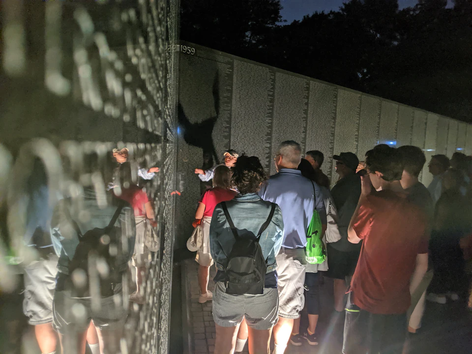 People gather around a reflective wall inscribed with names, illuminated at night. A guide gestures towards the inscriptions.