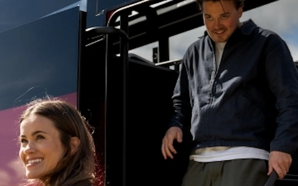 A group of four people smiling as they exit a red bus with windows, set outdoors under a partly cloudy sky.
