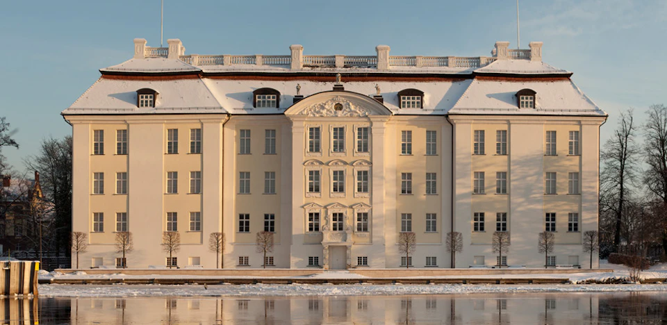 A large, cream-colored, symmetrical building with a decorative façade, windows, and a snow-covered roof reflected in a still body of water.