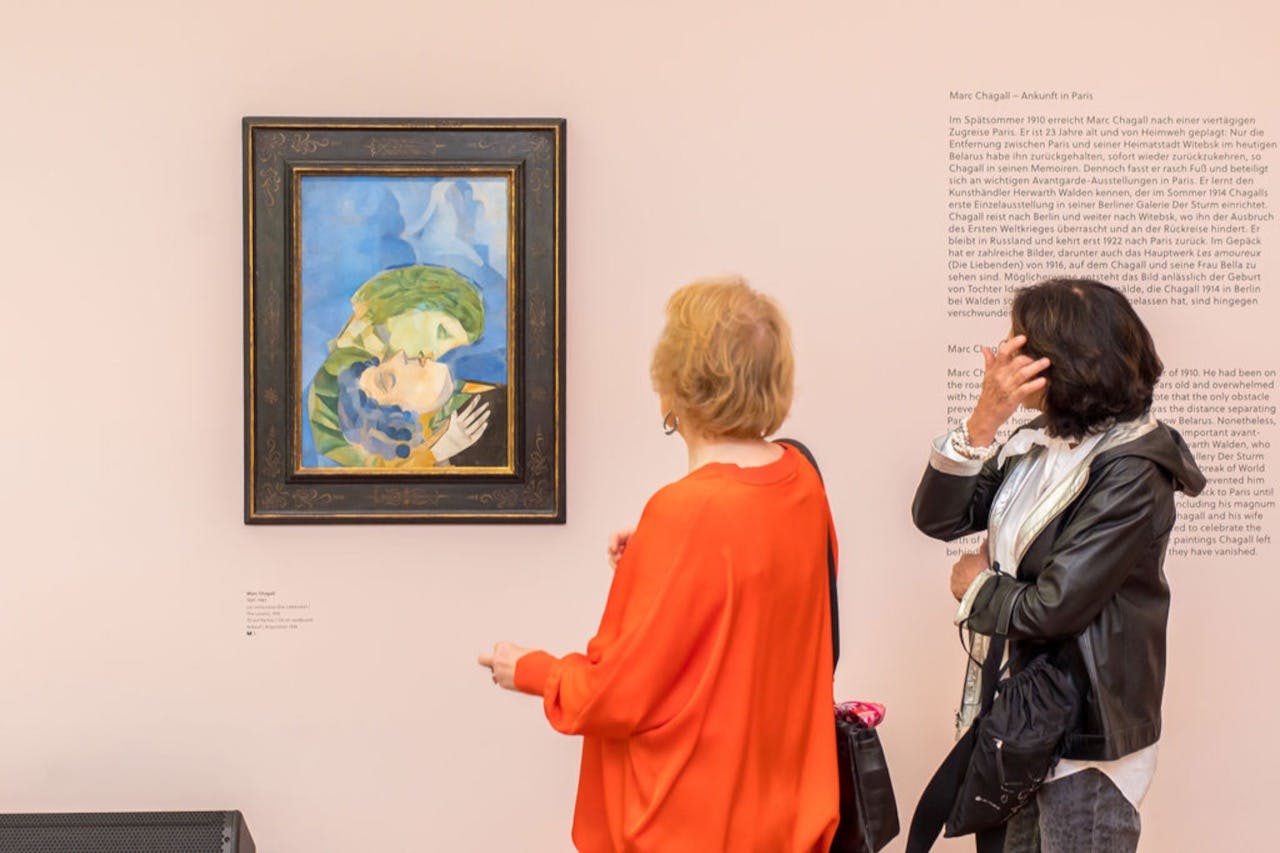 Dvě ženy pozorují zarámovaný obraz na zdi v galerii. Jedna žena nosí oranžový top a druhá černobílou bundu.