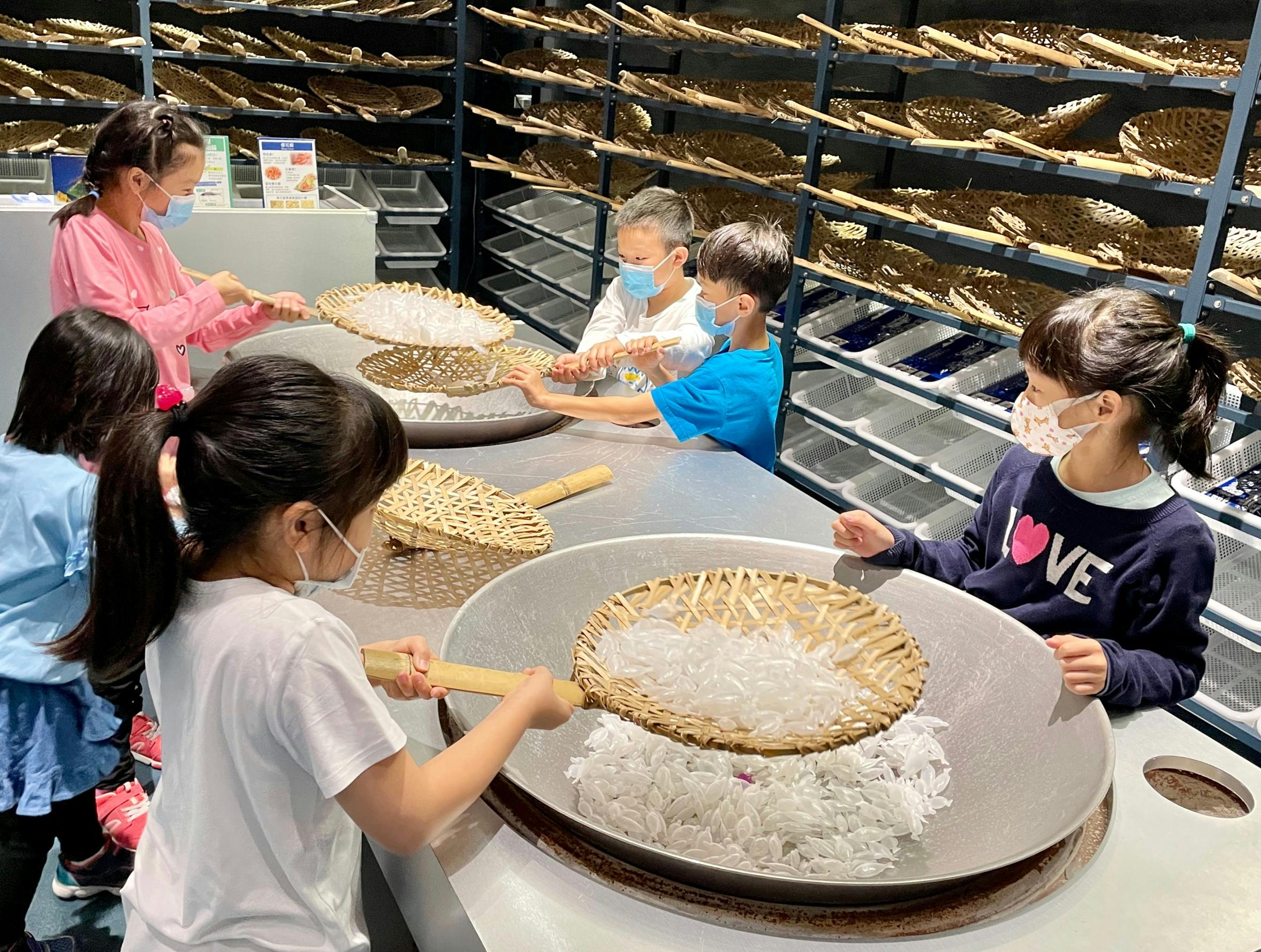 Children wearing masks use wicker baskets to handle white materials on a table in a workshop setting.