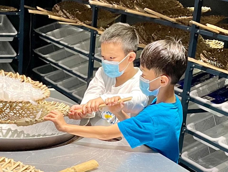 Children wearing masks are engaged in a hands-on activity with wicker baskets and trays, surrounded by drying racks.