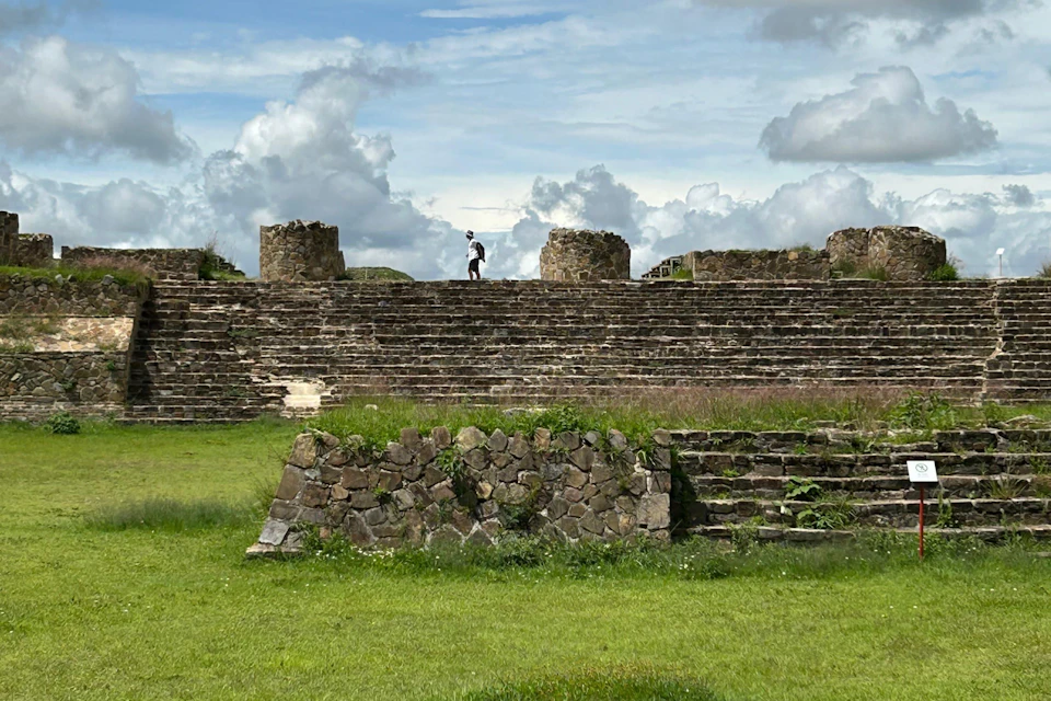 A person walks on top of an ancient stone step structure with grassy surroundings under a partly cloudy sky.
