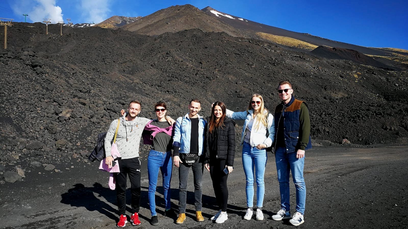 Vijf mensen staan voor een rotsachtig, vulkanisch landschap met een berg en schaarse sneeuwvlokken op de achtergrond.