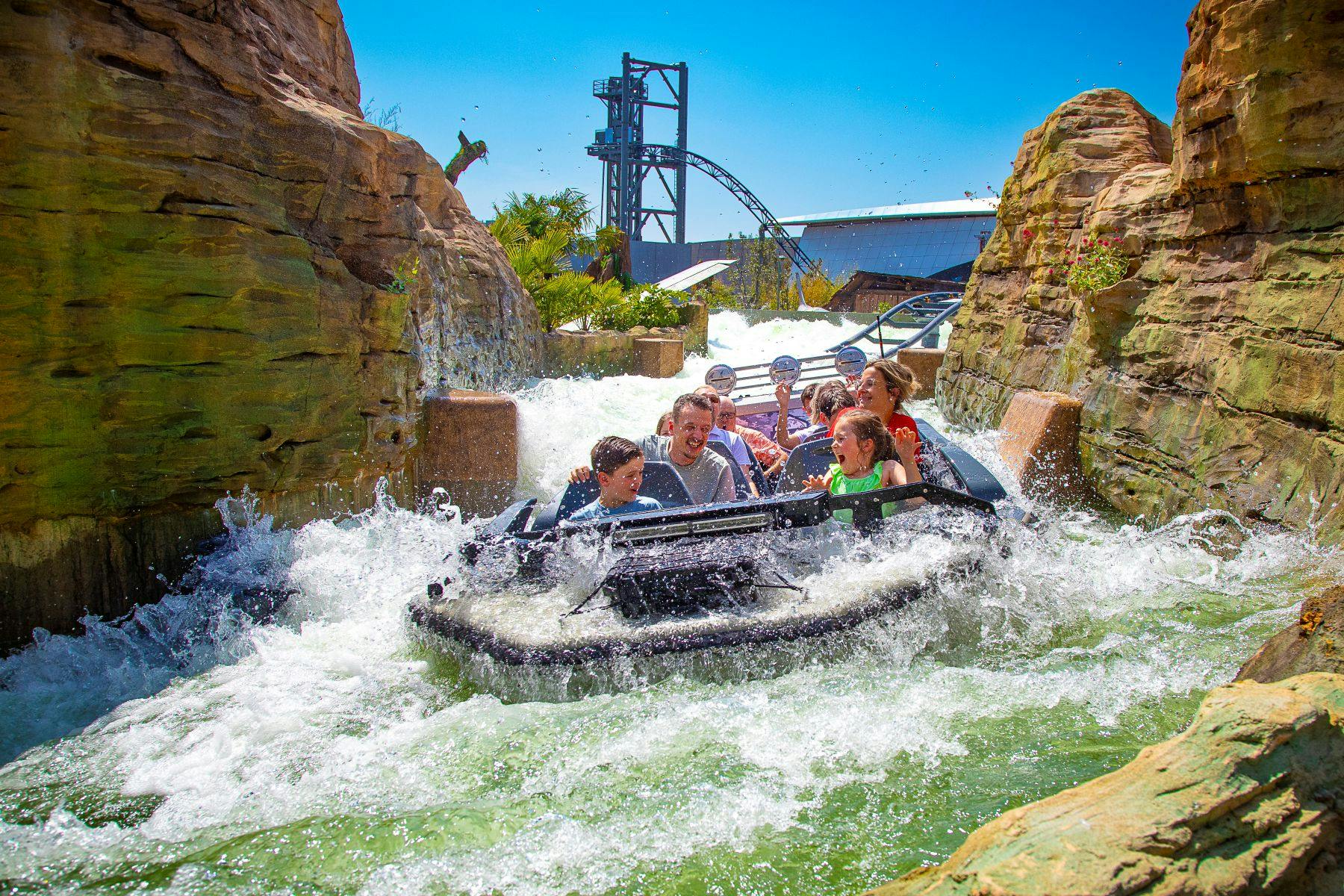 People enjoying a water ride, with splashes and rapids between rock formations under a sunny blue sky. Rollercoaster seen in background.