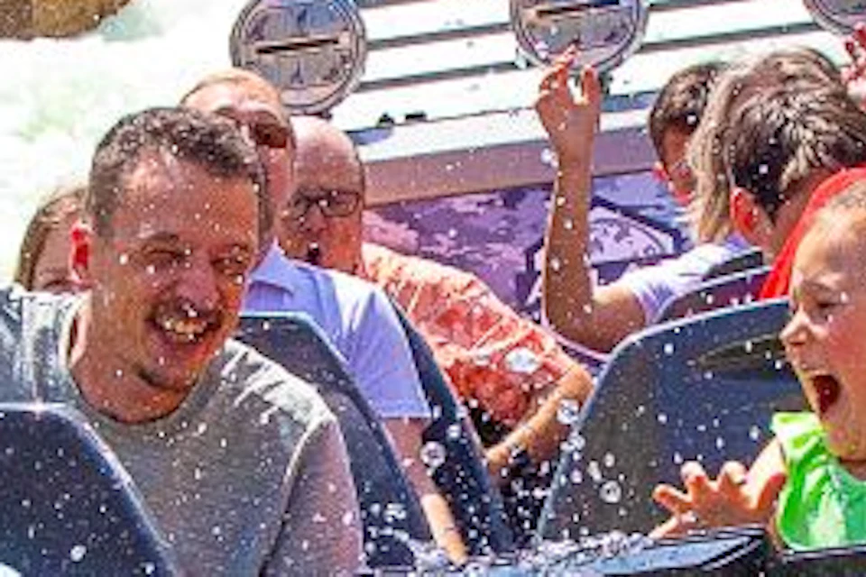 People enjoying a water ride, with splashes and rapids between rock formations under a sunny blue sky. Rollercoaster seen in background.