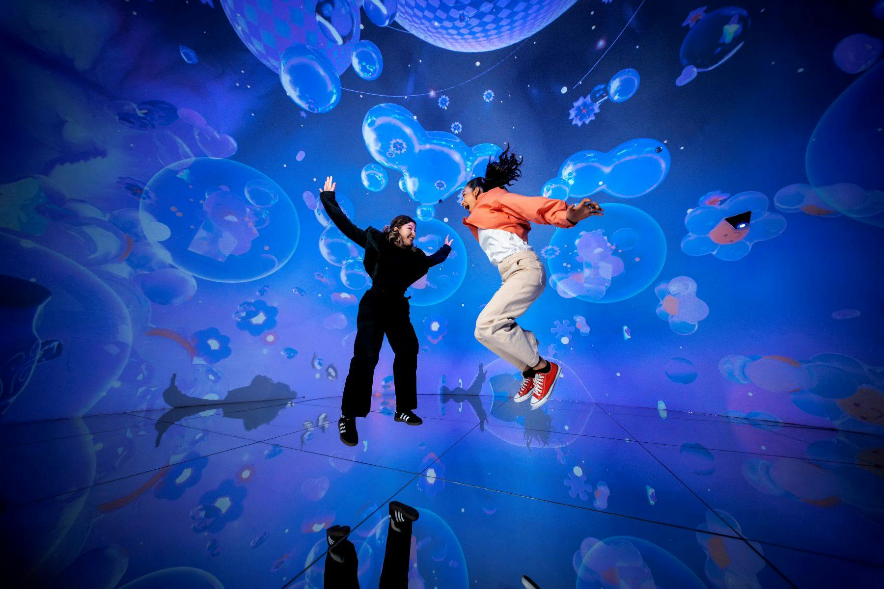 Zwei Menschen springen freudig in einer lebendigen, immersiven Umgebung mit bunten Blasen und schimmernden Lichtern um sie herum.